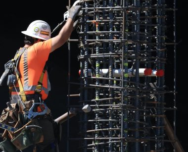 Worker in safety gear climbs a steel scaffolding structure on a construction site under daylight.